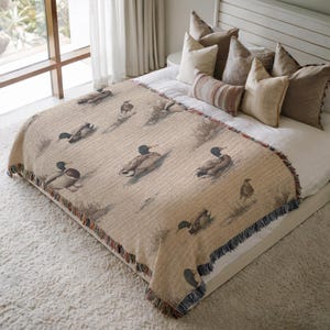 May include: A beige throw blanket with a duck pattern. The blanket features multiple ducks in a natural setting, with a fringed edge in shades of blue, red, and brown. The blanket is on a bed with several pillows.