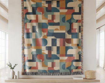 May include: A woven throw blanket featuring a geometric pattern of interlocking shapes in shades of navy blue, terracotta orange, muted teal, and cream. The rectangular blanket has a fringed edge along the top and bottom borders.
