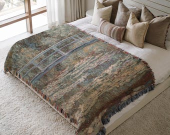 Op de afbeelding: Een geweven sprei met een brugontwerp in groen, blauw en bruin. De deken, met een gefranjerde rand, is over een bed gedrapeerd. Het kunstwerk op de deken lijkt op een aquarel.