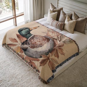 May include: Beige throw blanket featuring a detailed watercolor painting of a mallard duck surrounded by autumn leaves. The blanket has a fringed edge and is displayed on a bed with neutral-toned pillows.