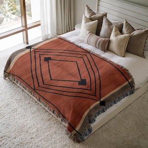 Op de afbeelding: Een terracotta kleurige sprei met een geometrisch ruitpatroon in zwart. De sprei heeft een gefranjerde rand en wordt getoond op een bed met wit beddengoed en decoratieve kussens. De sprei meet ongeveer 152 cm bij 127 cm.