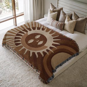 Puede incluir: Una manta tejida con un diseño de sol con una cara en tonos marrones, beige y crema. La manta tiene un borde con flecos y está colocada sobre una cama. La manta mide aproximadamente 152 cm por 127 cm.