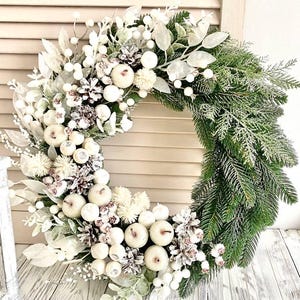 May include: A festive winter wreath featuring a mix of white and silver accents. The wreath is adorned with white berries, pinecones, and faux pumpkins, set against a backdrop of green pine needles. The wreath is displayed on a white wooden surface.