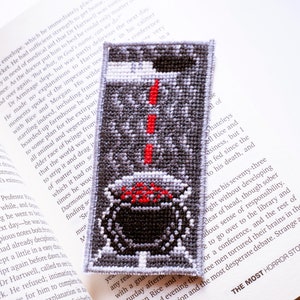 May include: A black and white cross-stitch bookmark featuring a cauldron with red flames and a black cat. The bookmark is on a book with text about a horror story.