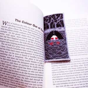 K&ouml;nnte beinhalten: Nahaufnahme eines Buches mit dem Titel "The Colour Out of Space" und einem Kreuzstich-Lesezeichen, das einen dunklen Wald mit einem roten und wei&szlig;en Haus darstellt. Der Text auf der Seite lautet "West of Arkham the hills rise wild, and there are narrow glens where the trees slope fantastically, and with deep woods that no axe has ever cur."