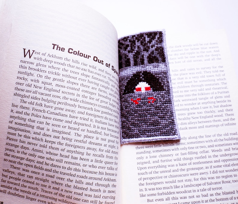 K&ouml;nnte beinhalten: Nahaufnahme eines Buches mit dem Titel "The Colour Out of Space" und einem Kreuzstich-Lesezeichen, das einen dunklen Wald mit einem roten und wei&szlig;en Haus darstellt. Der Text auf der Seite lautet "West of Arkham the hills rise wild, and there are narrow glens where the trees slope fantastically, and with deep woods that no axe has ever cur."
