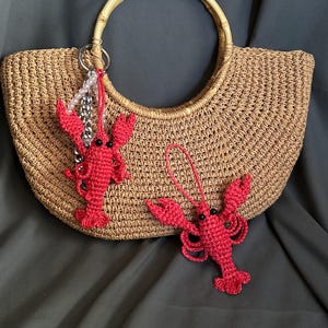 Puede incluir: Un bolso de mano de crochet marr&oacute;n con dos abalorios de langosta de crochet rojo. Un abalorio de langosta est&aacute; unido a una cadena plateada con cuentas blancas.