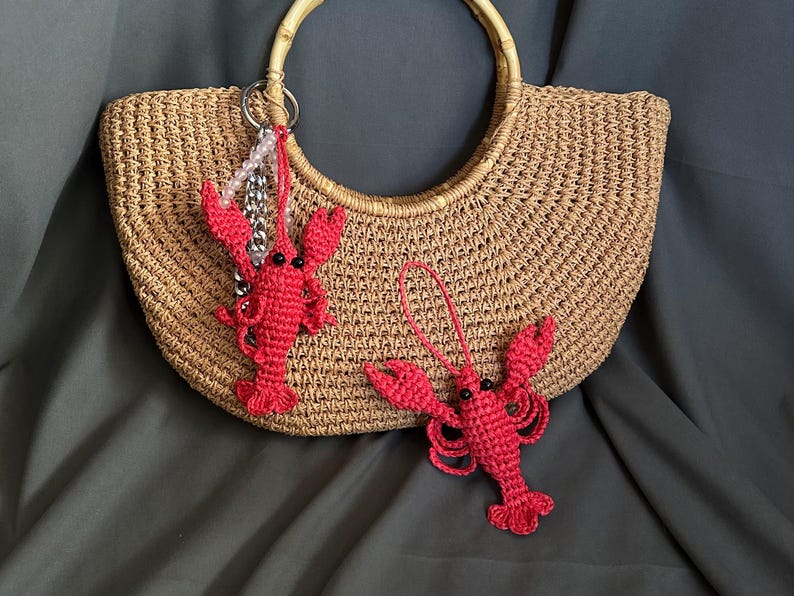 Puede incluir: Un bolso de mano de crochet marr&oacute;n con dos abalorios de langosta de crochet rojo. Un abalorio de langosta est&aacute; unido a una cadena plateada con cuentas blancas.