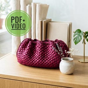 May include: A deep magenta, hand-crocheted clutch bag with a gathered top. The bag is displayed on a wooden surface, alongside a small white vase with dried flowers, and a brass plant stand with green leaves. The image includes the text "PDF + VIDEO".