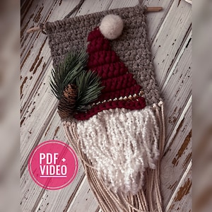 May include: A crocheted gnome wall hanging with a red hat, white beard, and a pine sprig. The gnome is hanging on a whitewashed wood background.