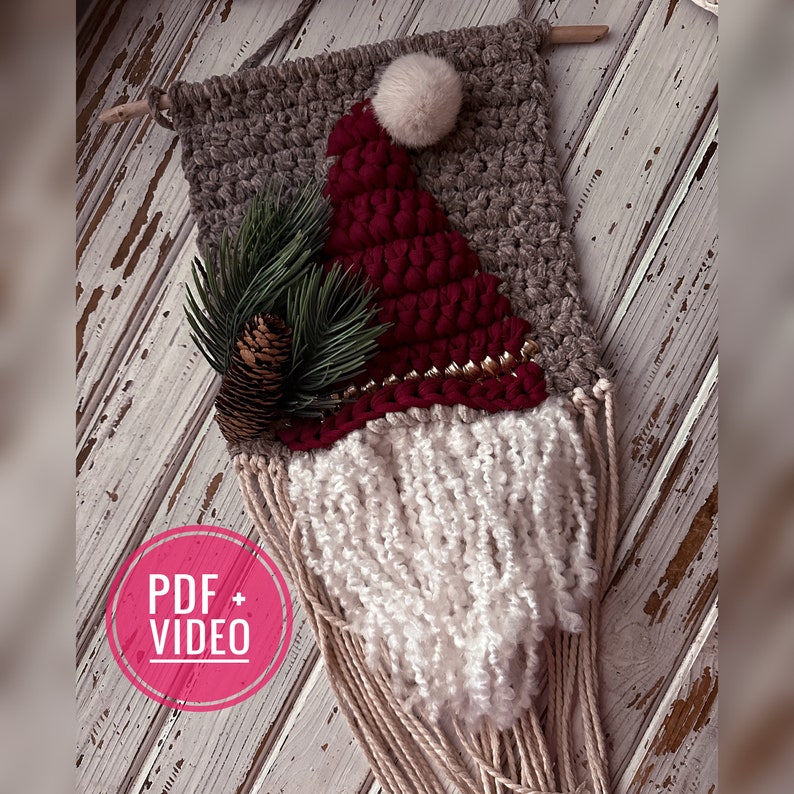 May include: A crocheted gnome wall hanging with a red hat, white beard, and a pine sprig. The gnome is hanging on a whitewashed wood background.