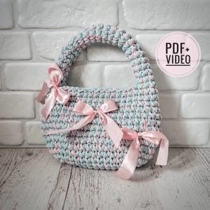 Pode incluir: Uma bolsa de crochê de cor azul claro e rosa. A bolsa tem uma alça e é decorada com fitas rosa.