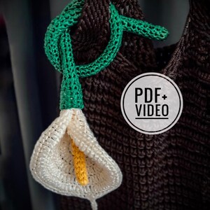 May include: A crocheted calla lily with a yellow stamen and a green stem and knot. The flower is white, and the background is a brown crocheted fabric. The image includes a circular graphic with the text "PDF+VIDEO".