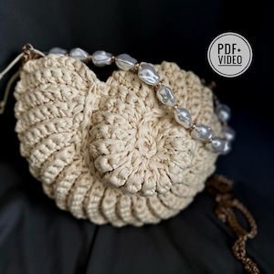 May include: A cream-colored, crocheted handbag with a circular design. The bag is adorned with a strand of large, irregular, off-white pearls. A gold-colored chain and a tag that says "PDF+VIDEO" are also visible.