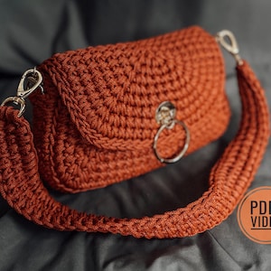 Può includere: Una borsa all'uncinetto color ruggine con chiusura a patta e una lunga tracolla. La borsa presenta un dettaglio ad anello color argento sul davanti e chiusure color argento. La tracolla è spessa e all'uncinetto.