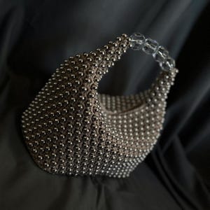 May include: A silver beaded handbag with a clear plastic handle. The bag is made of small, round beads that are tightly woven together.