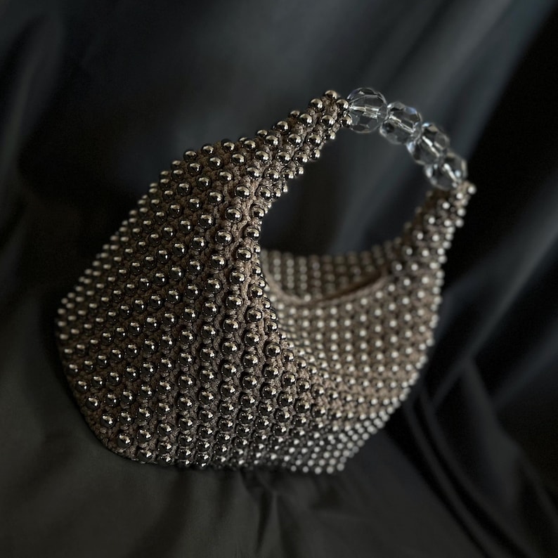 May include: A silver beaded handbag with a clear plastic handle. The bag is made of small, round beads that are tightly woven together.