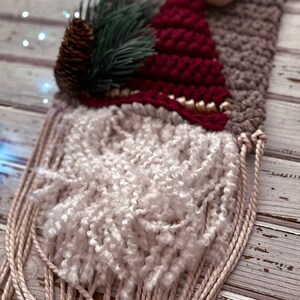 May include: A woven wall hanging featuring a gnome with a red hat, white beard, and a pine sprig. The gnome is woven with various textures and colors, including brown, white, and red.