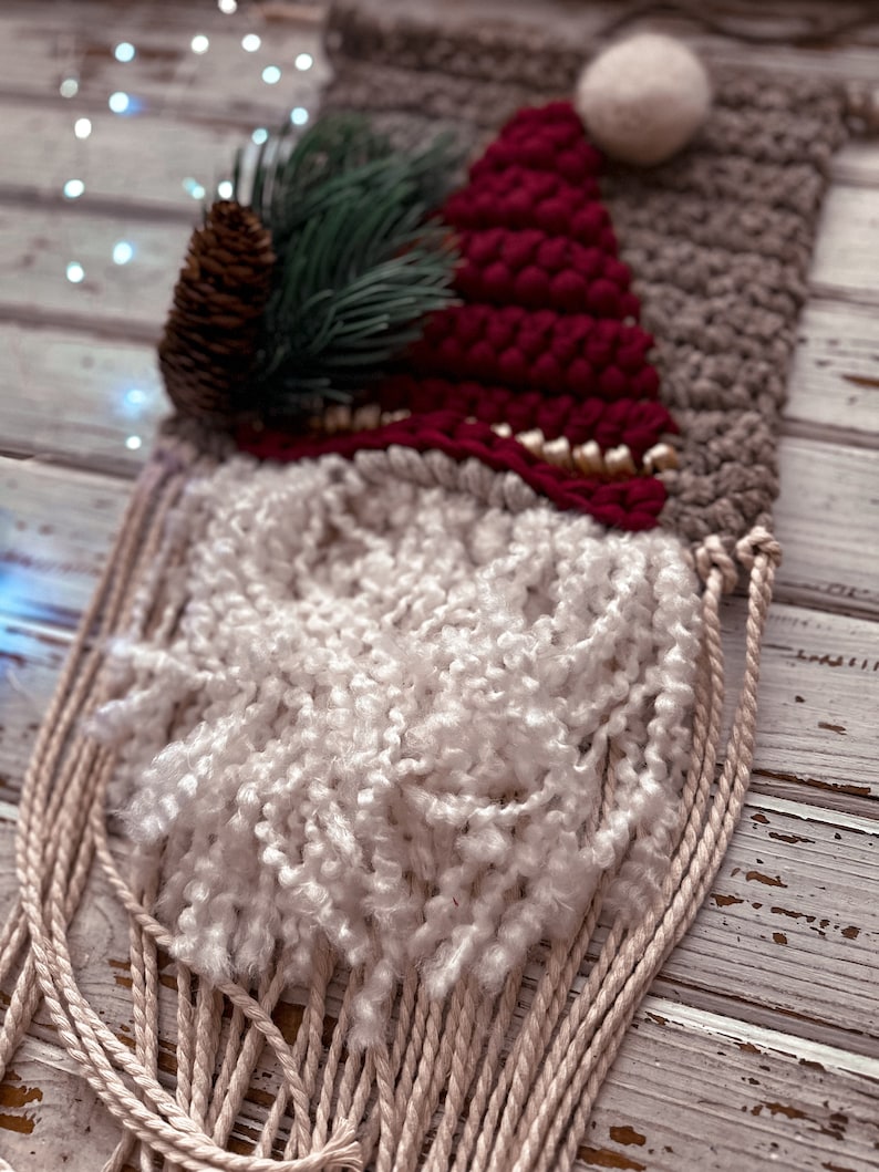 May include: A woven wall hanging featuring a gnome with a red hat, white beard, and a pine sprig. The gnome is woven with various textures and colors, including brown, white, and red.