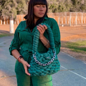 May include: A woman wearing a green jumpsuit holds a large, green knitted handbag with a silver chain strap.
