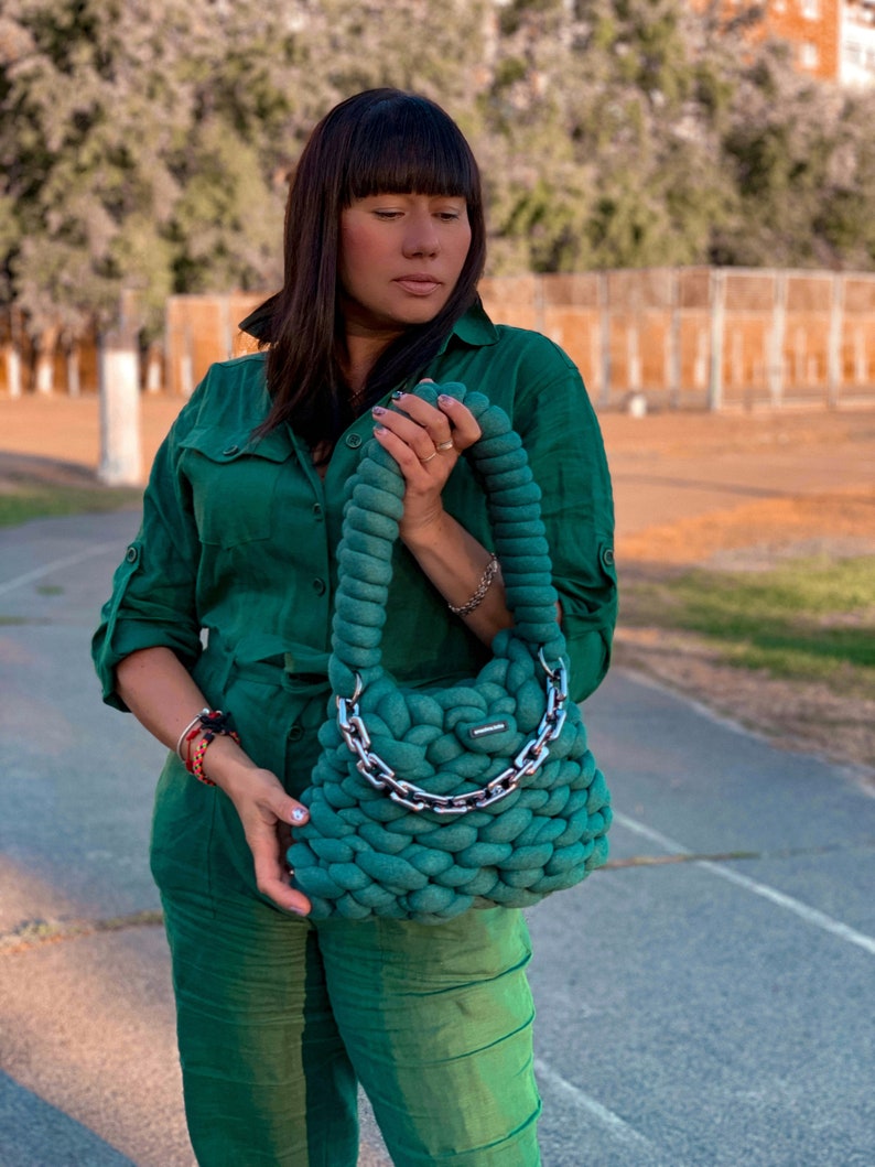 May include: A woman wearing a green jumpsuit holds a large, green knitted handbag with a silver chain strap.