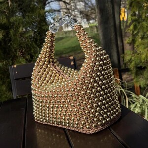 May include: A small, brown and gold beaded handbag with a clear plastic handle. The bag is made of crocheted beads and has a unique, textured design.