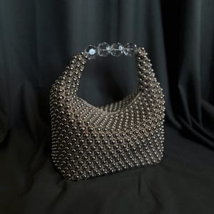 May include: A silver beaded handbag with a clear crystal handle. The bag is made of small, round beads that are woven together to create a textured surface.