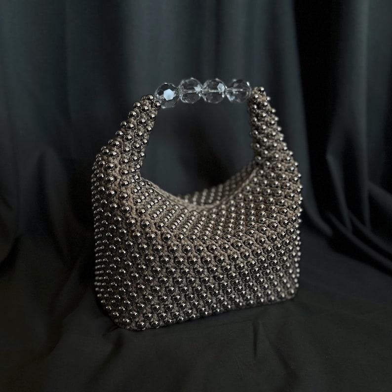 May include: A silver beaded handbag with a clear crystal handle. The bag is made of small, round beads that are woven together to create a textured surface.