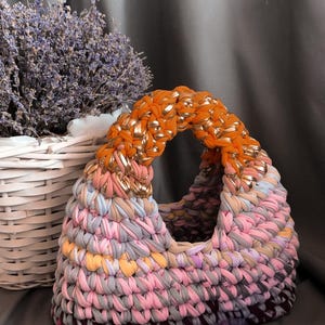 May include: A handmade, colorful tote bag with a rounded handle. The bag is made of knitted fabric in shades of pink, gray, orange, and gold. A white basket with lavender is in the background. The bag is a stylish accessory.