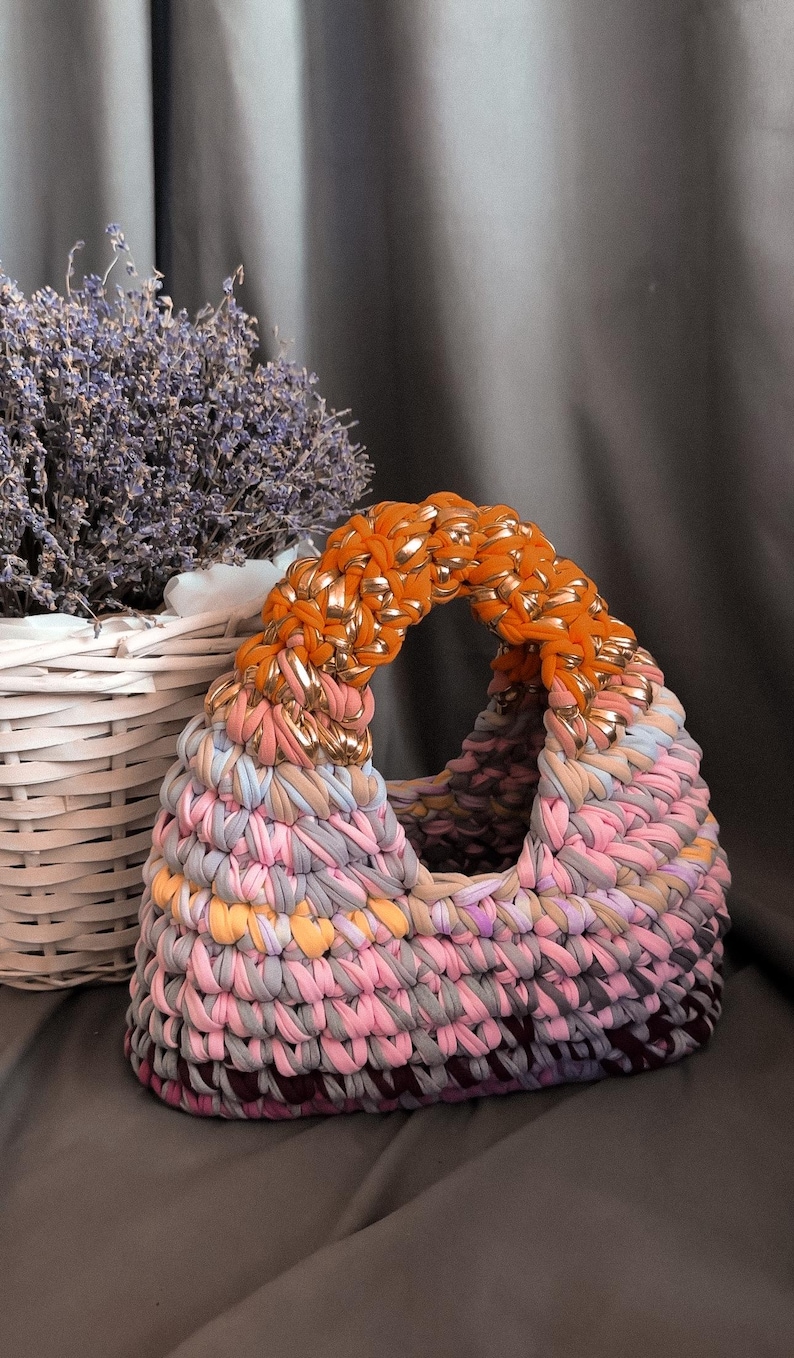 May include: A handmade, colorful tote bag with a rounded handle. The bag is made of knitted fabric in shades of pink, gray, orange, and gold. A white basket with lavender is in the background. The bag is a stylish accessory.