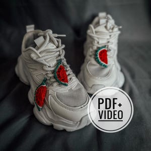 May include: White chunky sneakers with red and green watermelon slice charms hanging from the laces. The sneakers have a mesh and leather upper and a thick, textured sole. The image also includes a circular graphic with the text "PDF+VIDEO".