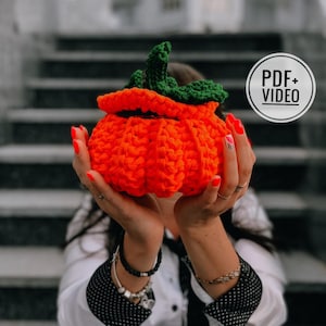 May include: A crocheted pumpkin-shaped container in orange with green leaves. The container is being held by a person's hands.