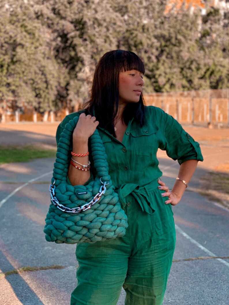 May include: A woman wearing a green jumpsuit and a green crocheted bag with a silver chain handle. The bag is worn over her shoulder.