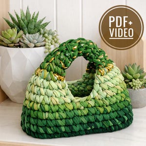 May include: A handmade green crochet bag with a rounded handle and gold accents. The bag is made of varying shades of green yarn, creating a textured appearance. The bag is displayed with succulent plants in white and gray pots.