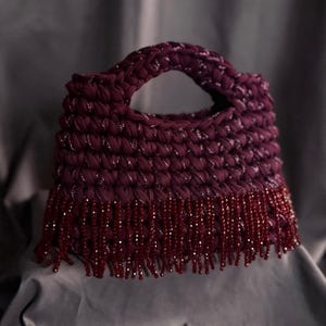 May include: A handmade, burgundy-colored handbag with a rounded handle. The bag is crocheted with a textured stitch and features silver metallic thread accents. The bottom edge is adorned with a fringe of burgundy beads, adding a decorative element.