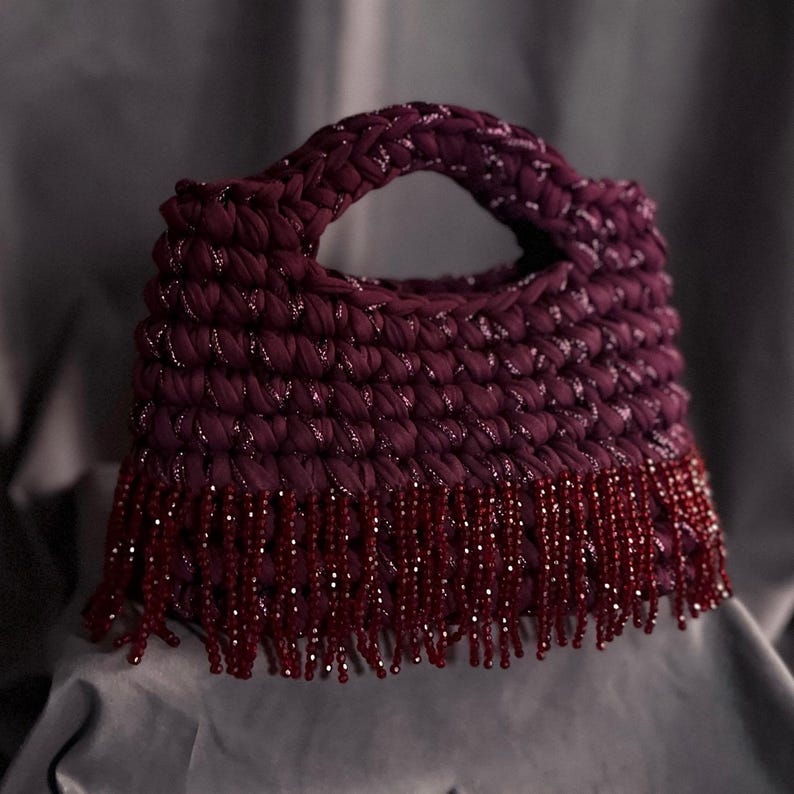 May include: A handmade, burgundy-colored handbag with a rounded handle. The bag is crocheted with a textured stitch and features silver metallic thread accents. The bottom edge is adorned with a fringe of burgundy beads, adding a decorative element.