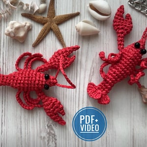 以下が含まれることがあります： 白い木製の背景に、赤い毛糸で編まれたロブスターの置物2つ。ロブスターは黒い目で、毛糸で編まれたハサミと尻尾が細部まで表現されている。背景には、ヒトデ、貝殻、銀色のチェーンがある。