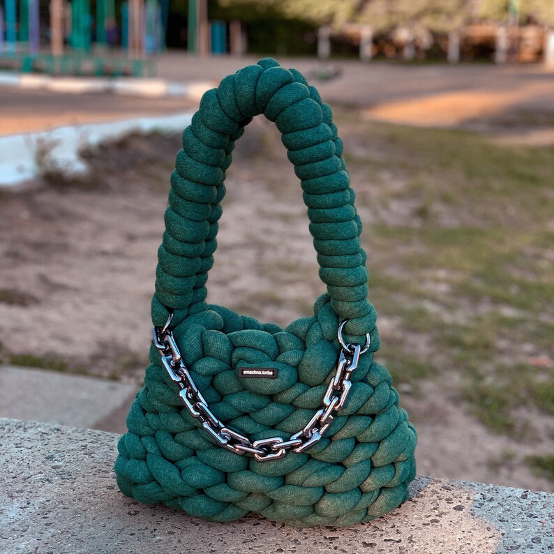 May include: A dark green crocheted tote bag with a silver chain strap. The bag has a small label that reads "handmade love".