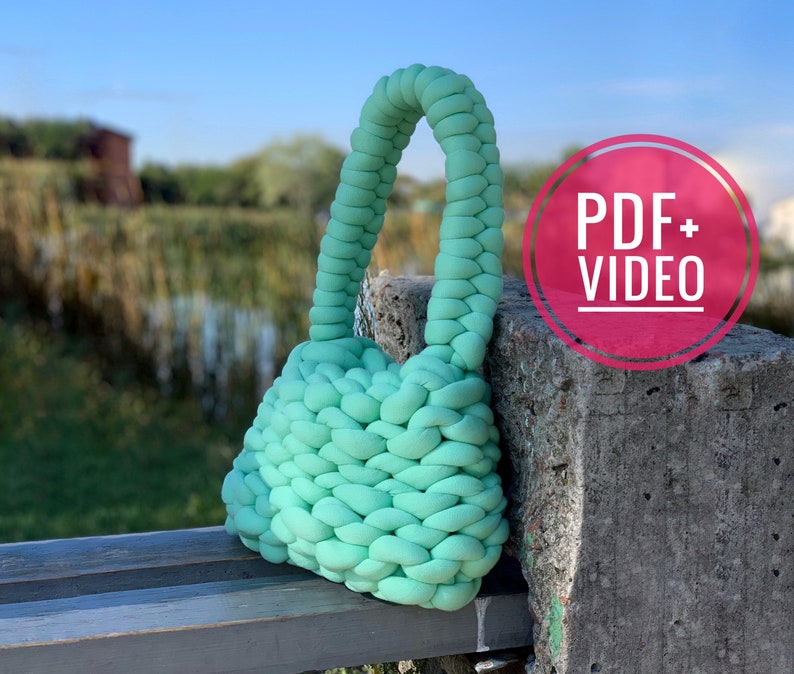 May include: A light green knitted bag with a thick rope handle. The bag is sitting on a wooden bench with a concrete block behind it. The text "PDF + VIDEO" is on a pink circle.