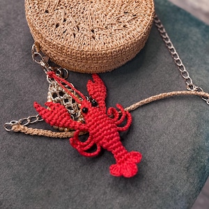 Puede incluir: Un amuleto de langosta rojo de ganchillo cuelga de un bolso tejido marr&oacute;n con una cadena plateada. El bolso y el amuleto est&aacute;n sobre una superficie de tela verde oscuro.