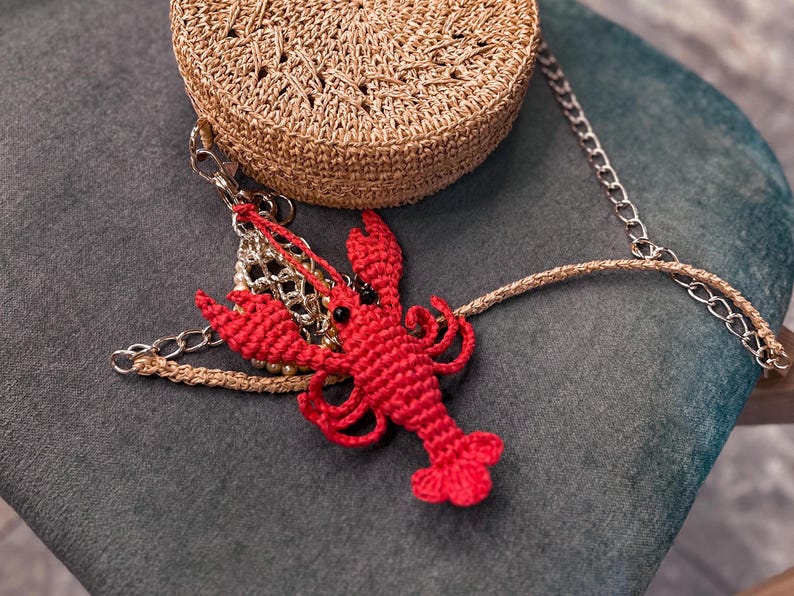 Puede incluir: Un amuleto de langosta rojo de ganchillo cuelga de un bolso tejido marr&oacute;n con una cadena plateada. El bolso y el amuleto est&aacute;n sobre una superficie de tela verde oscuro.