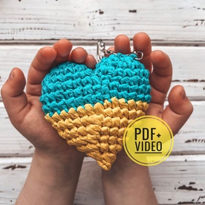 May include: A crocheted heart-shaped keychain in the colors of the Ukrainian flag, blue and yellow. The keychain is being held by a person's hands. The text "PDF + VIDEO" is on a yellow circle.