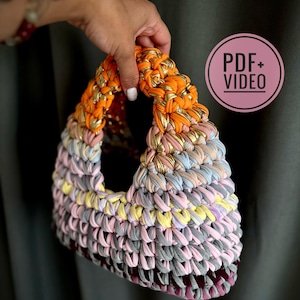 May include: A handmade, colorful crochet bag with an orange handle accented with gold metallic thread. The body of the bag features a mix of pastel colors, including pink, yellow, and gray. The bag is held by a hand.