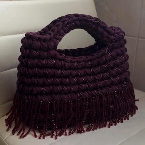 May include: A handmade, plum-colored handbag with a braided handle and a fringe of matching beads. The bag is made of a thick, textured material and sits on a white surface.