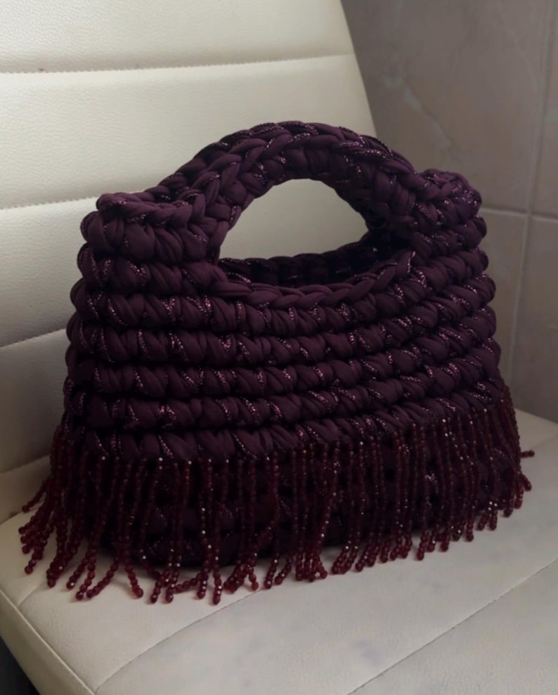 May include: A handmade, plum-colored handbag with a braided handle and a fringe of matching beads. The bag is made of a thick, textured material and sits on a white surface.