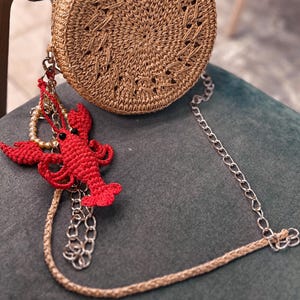 Puede incluir: Un bolso redondo de paja con un dise&ntilde;o de crochet y un dije de langosta unido a la correa de cadena.