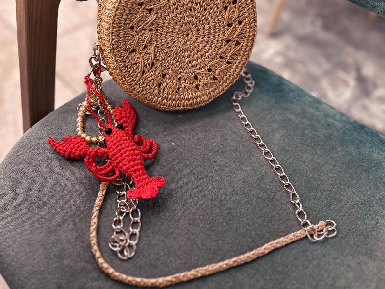 Puede incluir: Un bolso redondo de paja con un dise&ntilde;o de crochet y un dije de langosta unido a la correa de cadena.