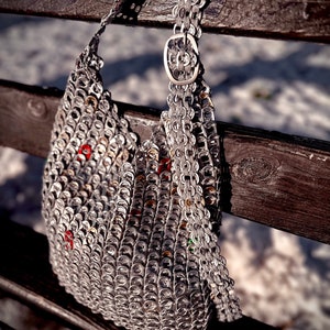 May include: A handmade silver shoulder bag constructed from recycled aluminum can tabs. The bag features a chain strap and a decorative buckle. The bag is resting on a wooden bench, with a snowy background.