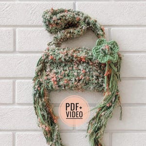 May include: A handmade crochet hat in shades of green and pink, with a flower accent. The hat has long, flowing strands and is displayed against a white brick wall. The image includes the text "PDF + VIDEO" in a circular graphic.