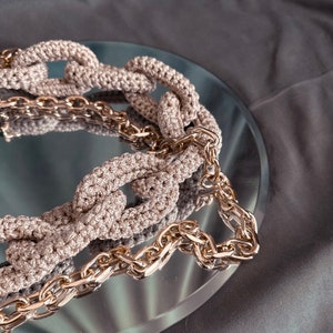 May include: A close-up of a beige crocheted chain necklace with a gold chain interwoven. The necklace is lying on a reflective surface.
